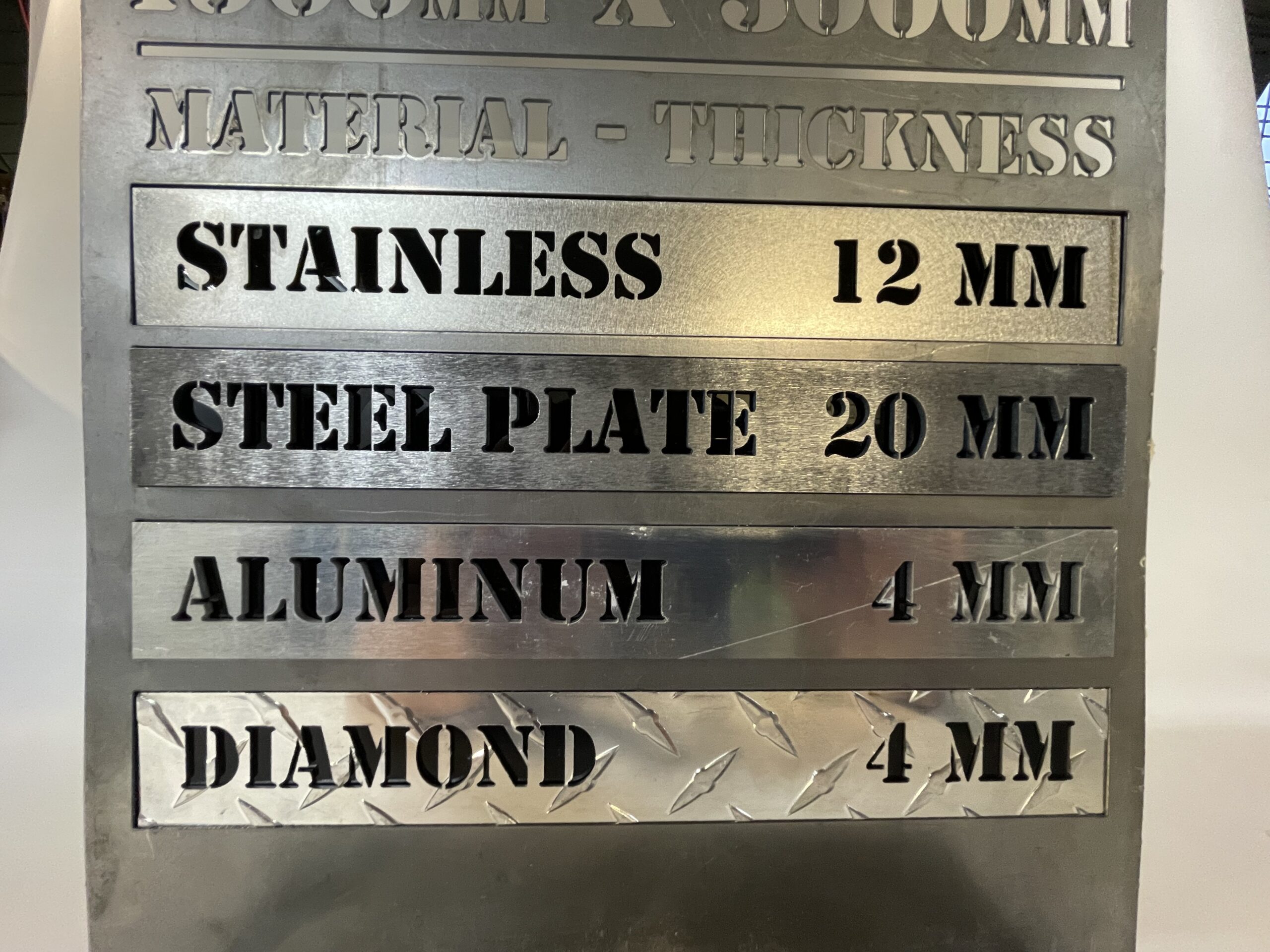 Engraved Metal Labels: laser cut sign with cut-out lettering listing different maximum material thicknesses for various metals. Stainless is 12 millimeters, Steel Plate is 20 millimeters, Aluminum is 4 millimeters, and Diamond is 4 millimeters.